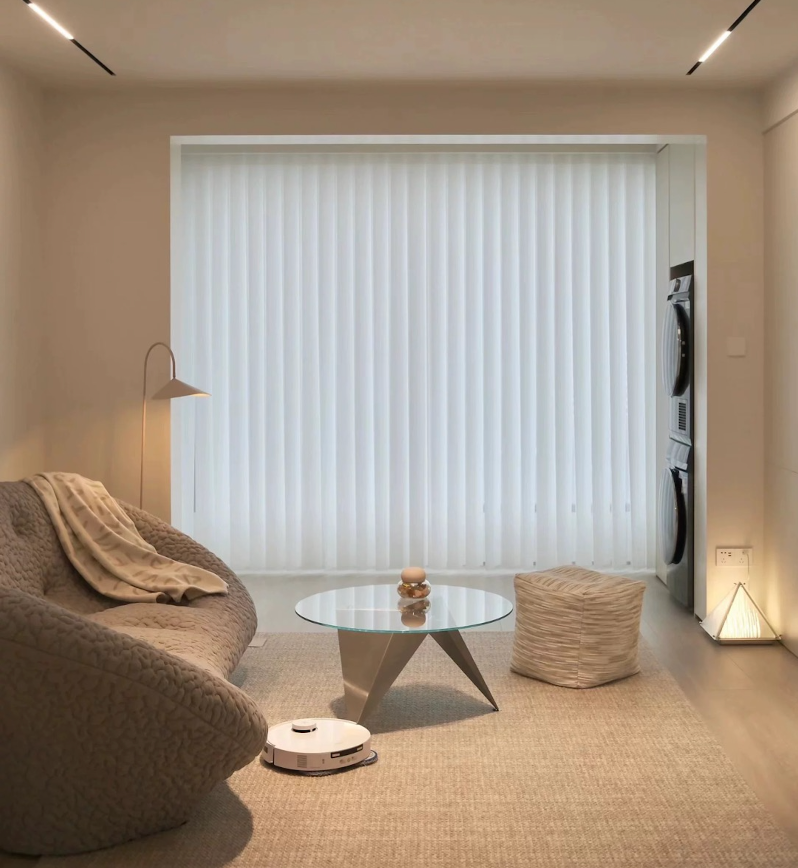 A white Ecovacs T30 Pro Omni robot vacuum cleaner operating in a modern, minimalistic living room with beige and neutral tones. The room features a cozy sofa with a blanket, a glass coffee table, a pouf, and large windows with vertical blinds