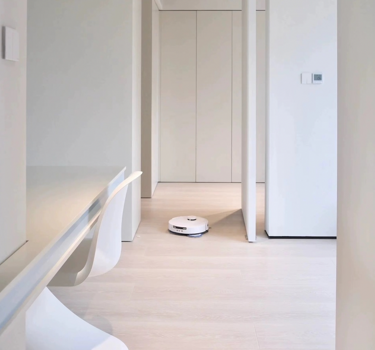 A white Ecovacs T30 Pro Omni robot vacuum cleaner navigating a modern, minimalistic home with light-colored hardwood floors. The space features clean lines, a white desk with a chair, and an open doorway leading to another room