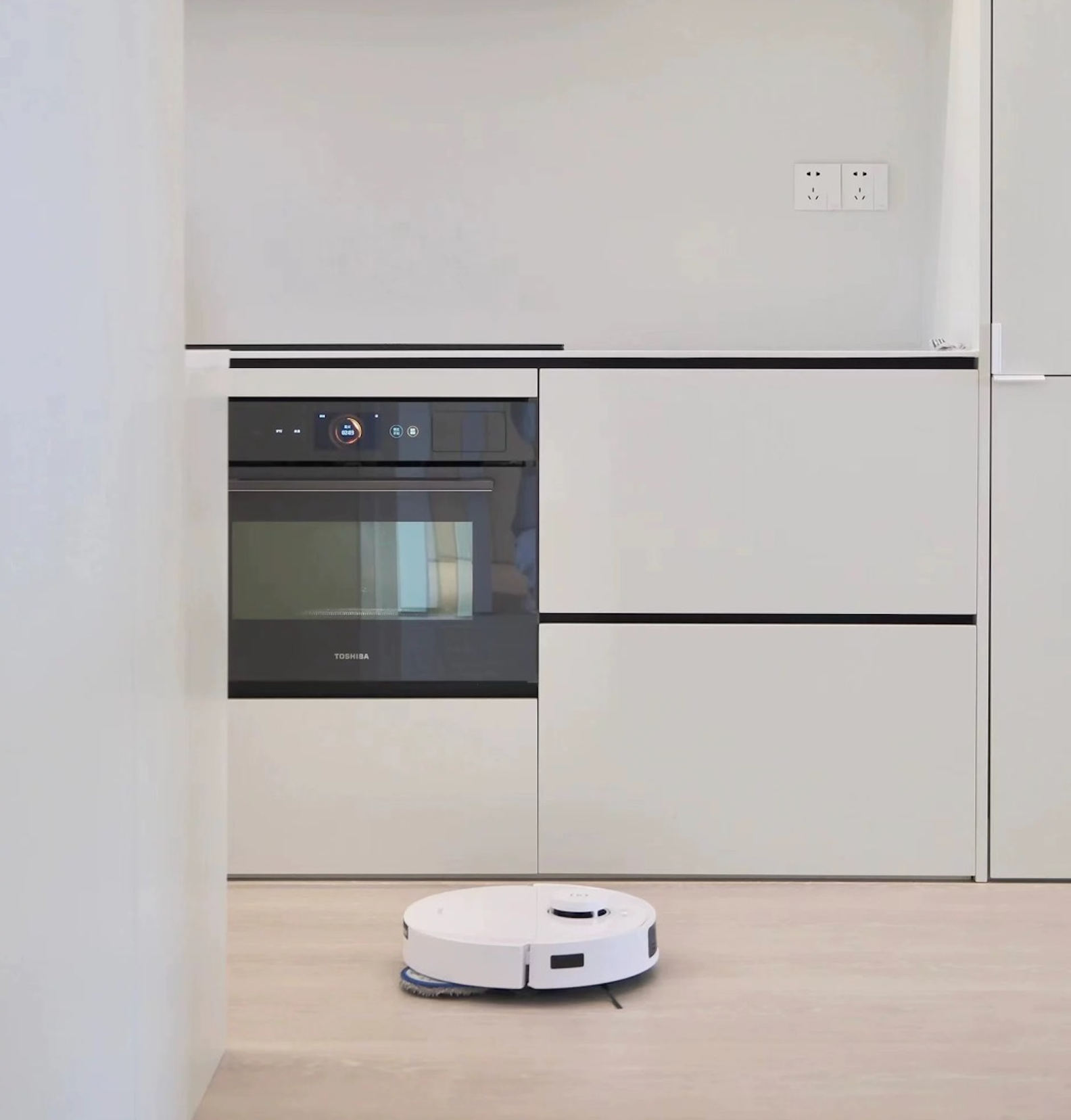 A white Ecovacs T30 Pro Omni robot vacuum cleaner operating in a modern kitchen with light-colored hardwood floors. The kitchen features sleek, minimalistic cabinetry and a built-in black and silver Toshiba oven