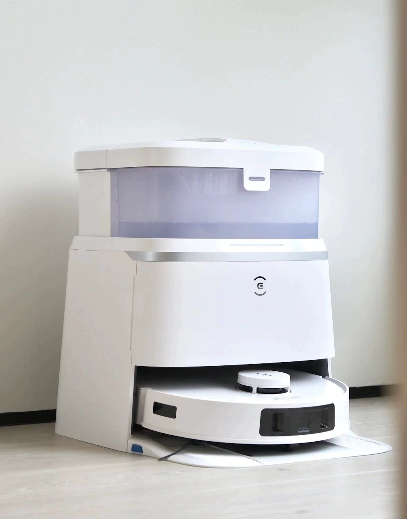 A white Ecovacs Deebot T30 Pro Omni robot vacuum cleaner docked in its charging and cleaning station. The station has a translucent water tank on top, and the vacuum's front sensors are visible. The setup is placed against a wall on a light-colored floor.